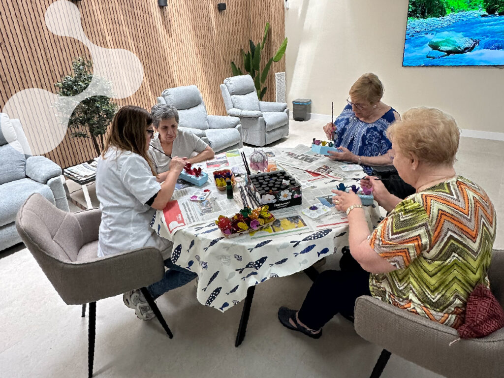 Grupo de personas mayores realizando una actividad manual de pintura en una sala luminosa, acompañadas por una persona que las asiste.