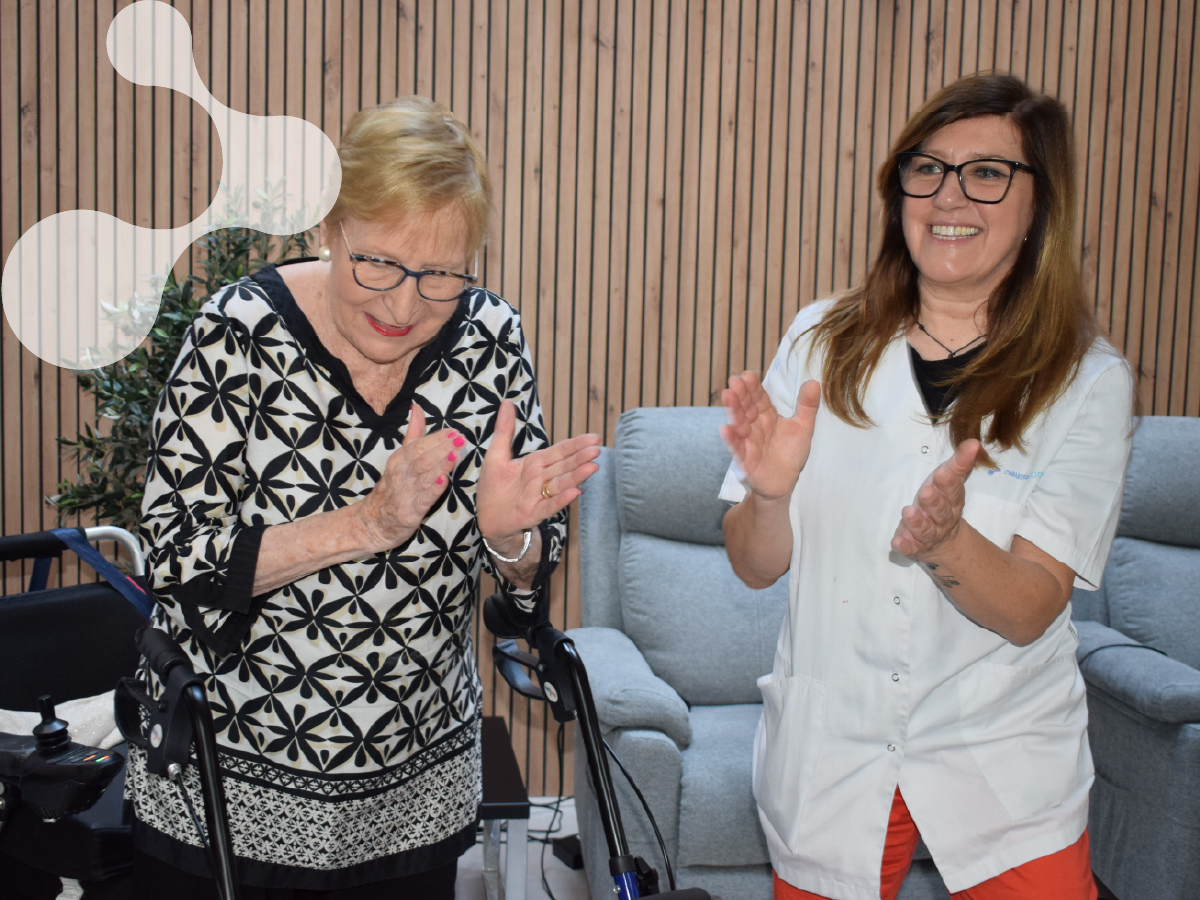 Una mujer mayor y una cuidadora sonríen y aplauden juntas en una sala luminosa y acogedora.